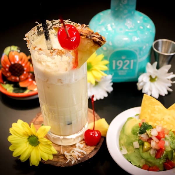 A pina colada with pineapple and cherry, guacamole with chips, and a blue tequila bottle.
