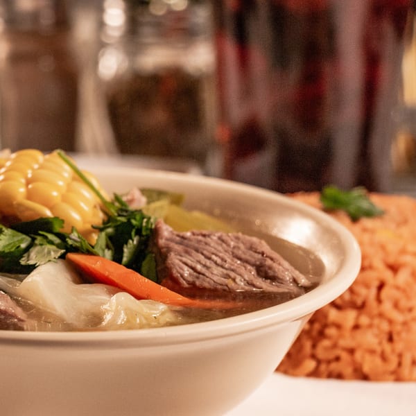 Hearty beef soup with corn, carrots, and cilantro, served with a side of red rice.