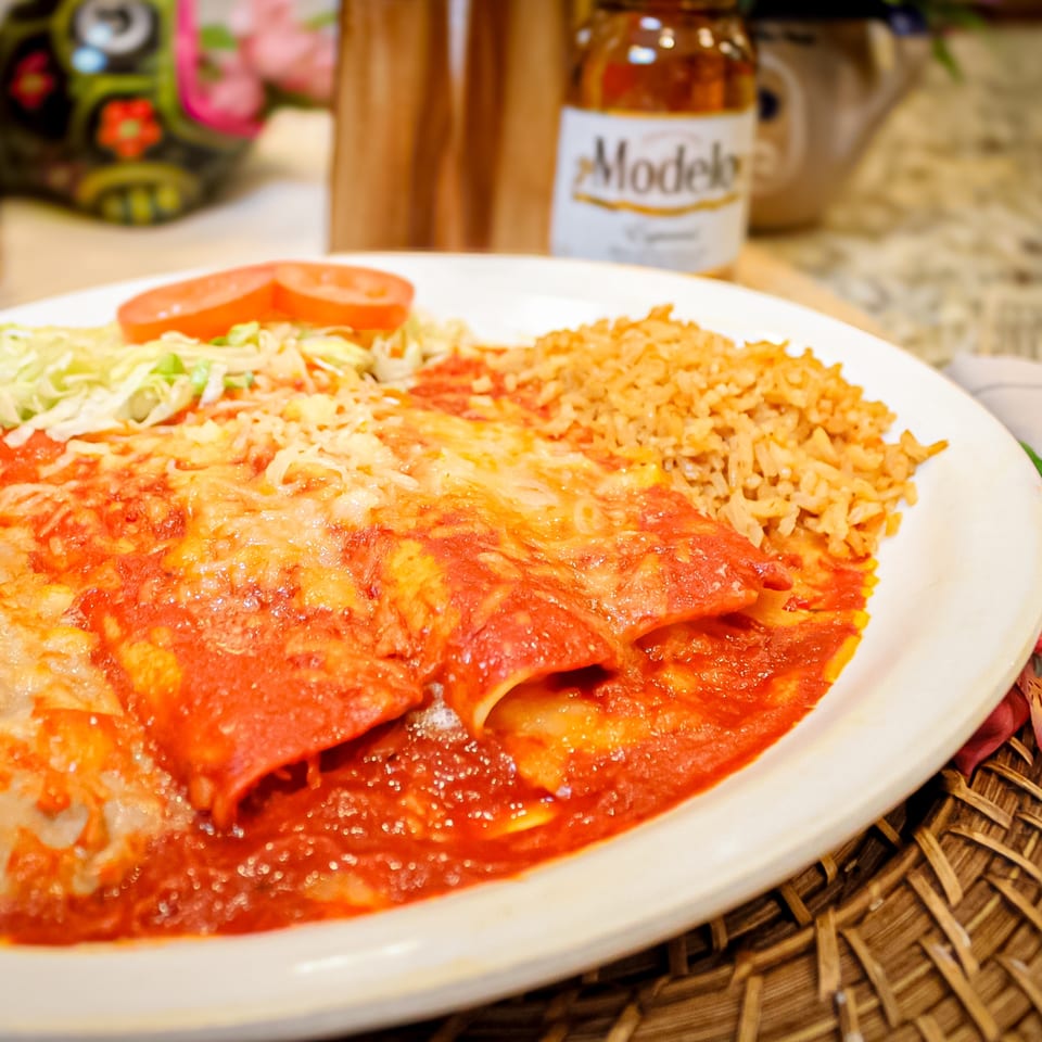 Cheese-covered enchiladas smothered in red sauce served with Mexican rice, lettuce, and tomato on a white plate.