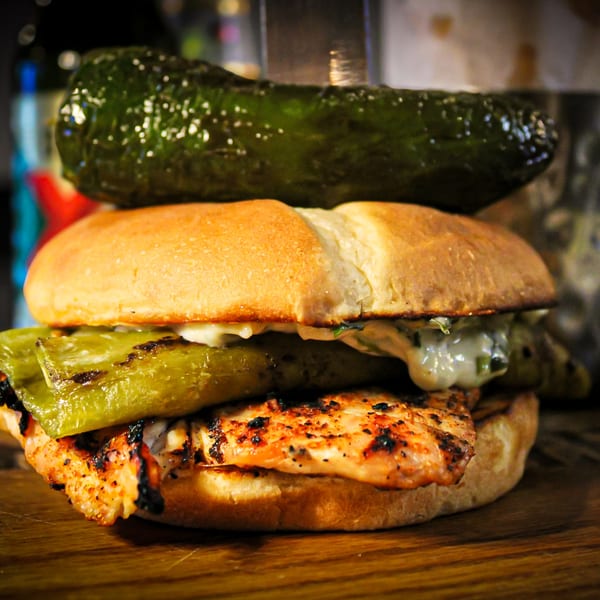 Grilled chicken sandwich with green peppers and a large pickle on top, served on a wooden board.