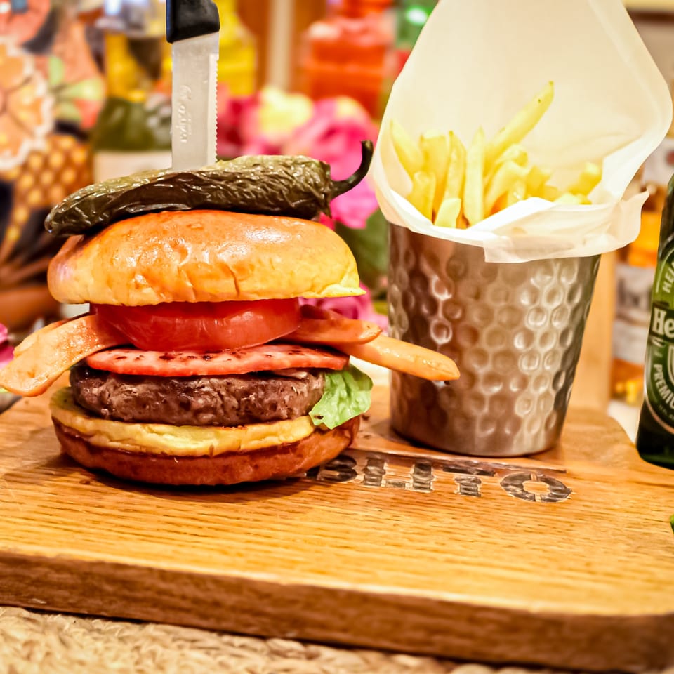 Gourmet burger topped with grilled pepper, tomato, bacon, and lettuce beside fries in a metal cup on a wooden table.