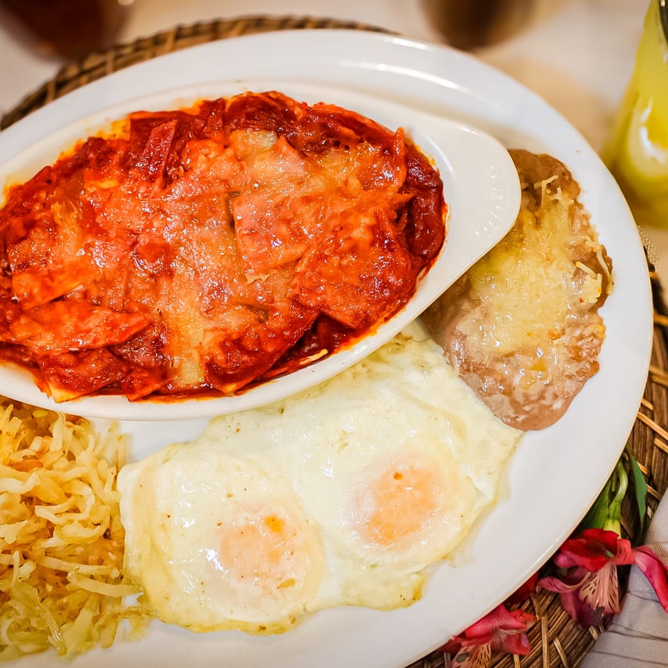 Plate of red-sauced pasta, two fried eggs, rice, and a breaded cutlet served with a yellow drink.