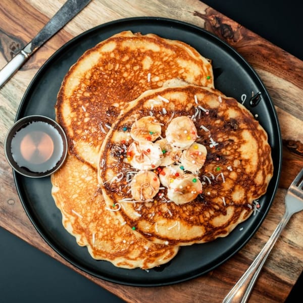Golden pancakes topped with sliced bananas, sprinkles, and coconut flakes, with a side of syrup.