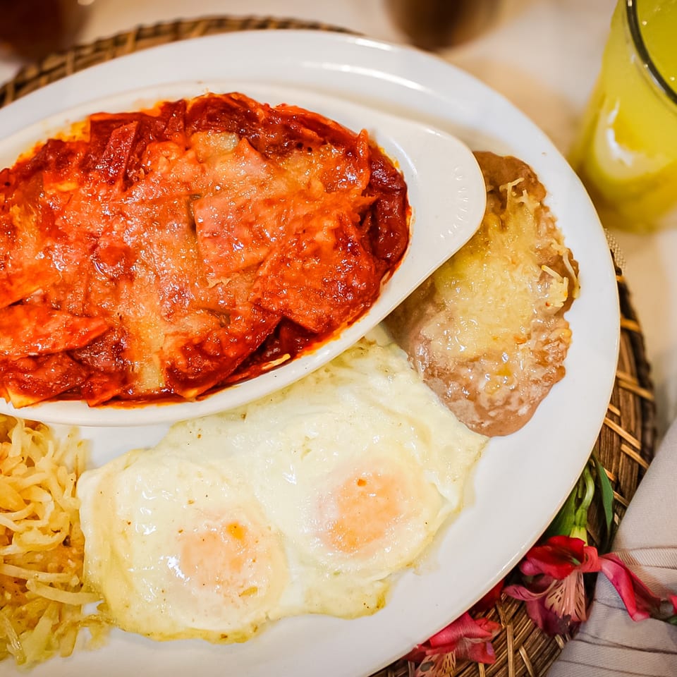 Plate of saucy enchiladas, fried eggs, rice, and beans served with a yellow beverage.