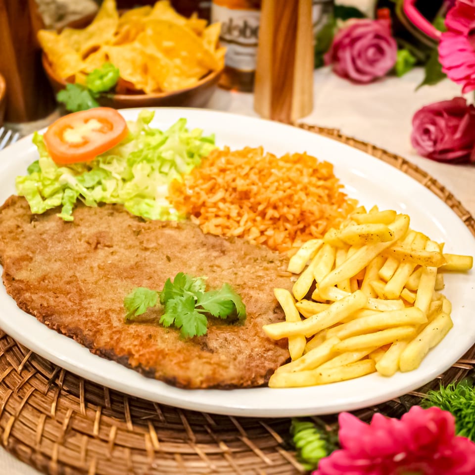Breaded steak served with French fries, seasoned rice, lettuce, tomato, and cilantro on a white plate.