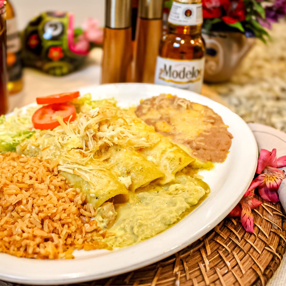 Mexican platter with enchiladas, rice, beans, salad, and tomato garnish beside a Modelo beer bottle.