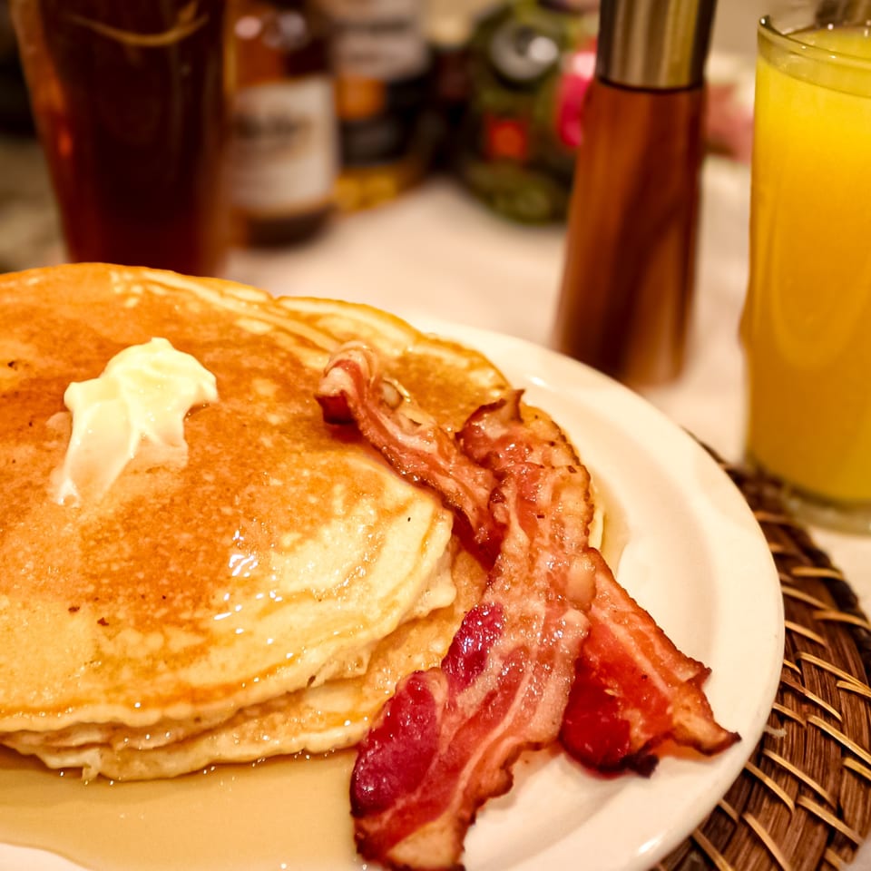 Stack of pancakes topped with butter and syrup, served with crispy bacon and orange juice.