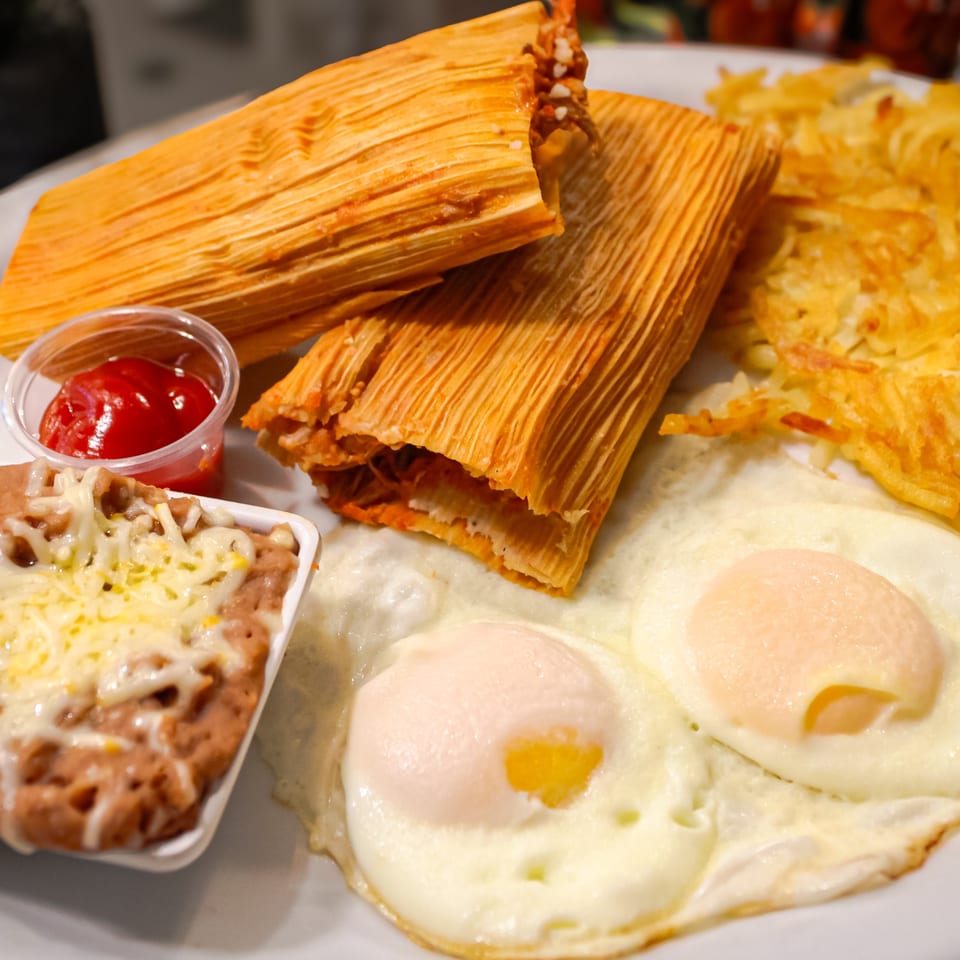 Mexican breakfast spread featuring husk-wrapped tamales, fried eggs, cheesy refried beans, hash browns, and ketchup.